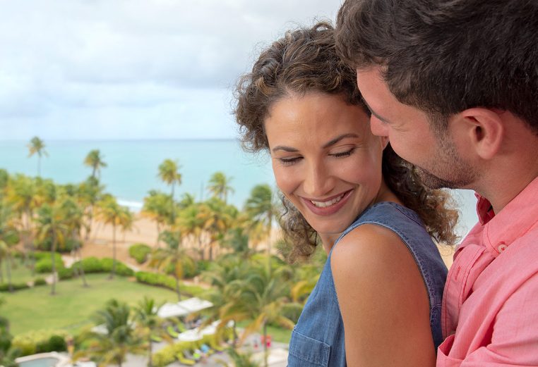 Man and woman smile on the balcony of their Margaritaville Vacation Club suite, with palm trees and ocean in the background.
